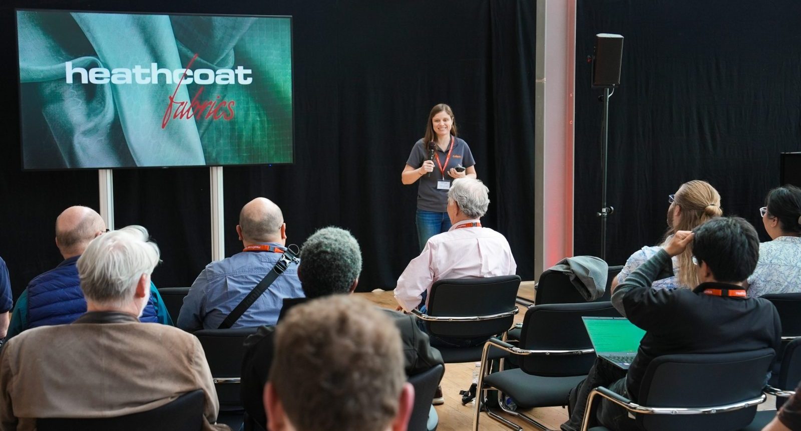 A woman speaks to a seated crowd of people at an exhibition
